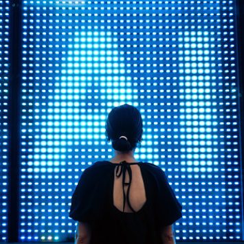 Rear view of young Asian businesswoman standing against illuminated digital display in city, using screen of AI technology. Concept of VR and AR technologies. Metaverse and futuristic data. People, AI, innovation and technology